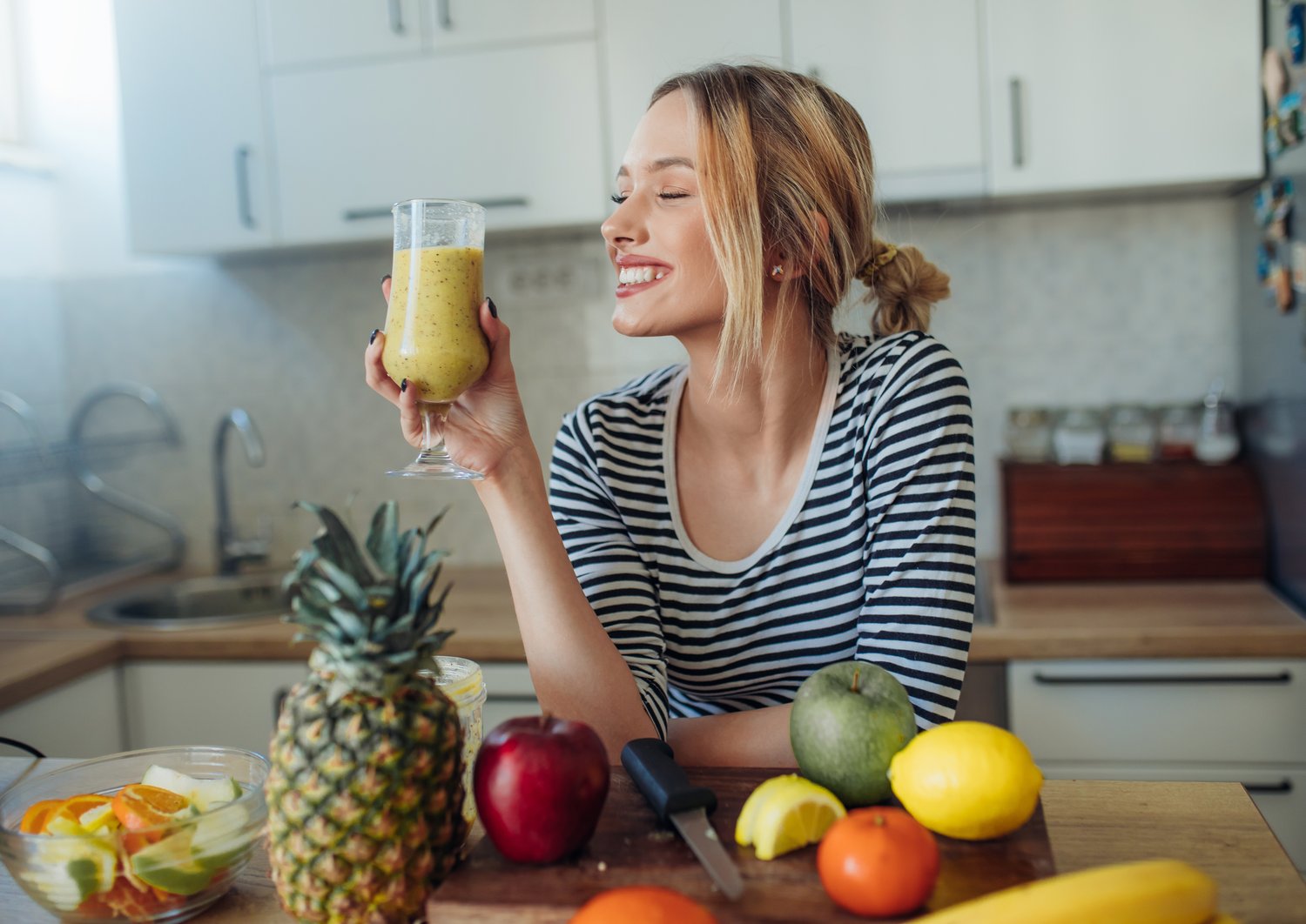 Frau mit gelbem Smoothie in der Küche. Vor ihr liegt verschiedenen Obst und Gemüse auf der Arbeitsplatte. 