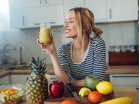 Frau mit gelbem Smoothie in der Küche. Vor ihr liegt verschiedenen Obst und Gemüse auf der Arbeitsplatte. 