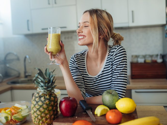 Frau mit gelbem Smoothie in der Küche. Vor ihr liegt verschiedenen Obst und Gemüse auf der Arbeitsplatte. 