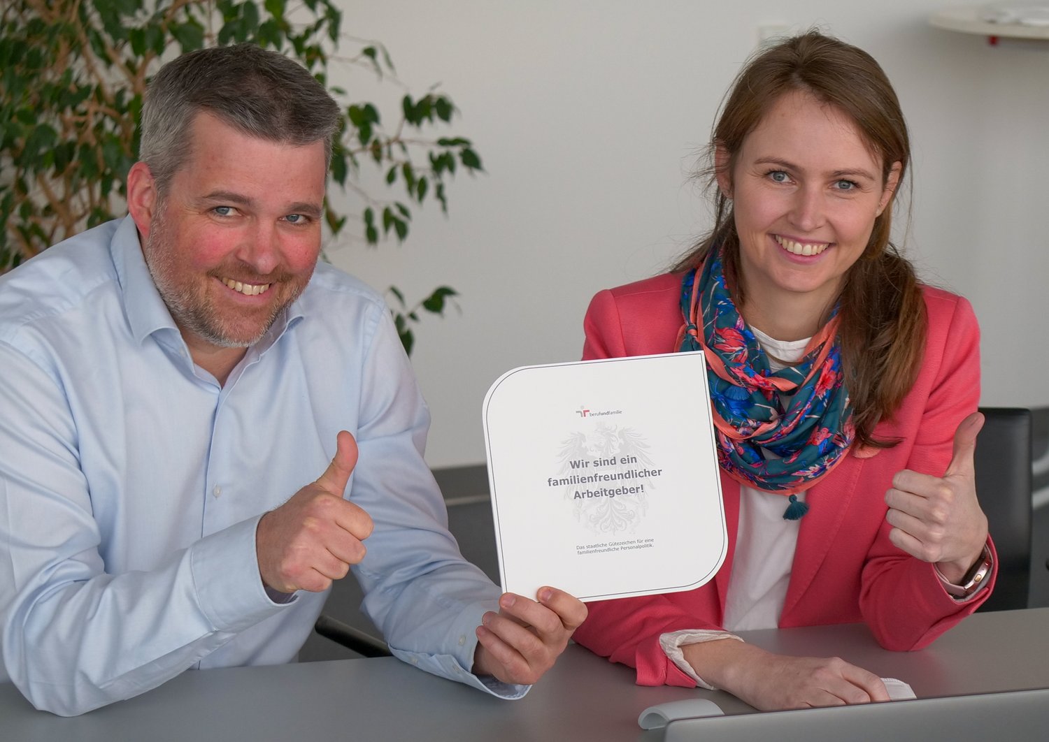 Geschäftsführer Gerald Murauer und Emily Knes, Head of Human Resources, freuen sich über die neuerliche Auszeichnung als familienfreundlicher Arbeitgeber. Sie sitzen nebeineinander mit einem Zertifikat und zeigen die Daumen hoch