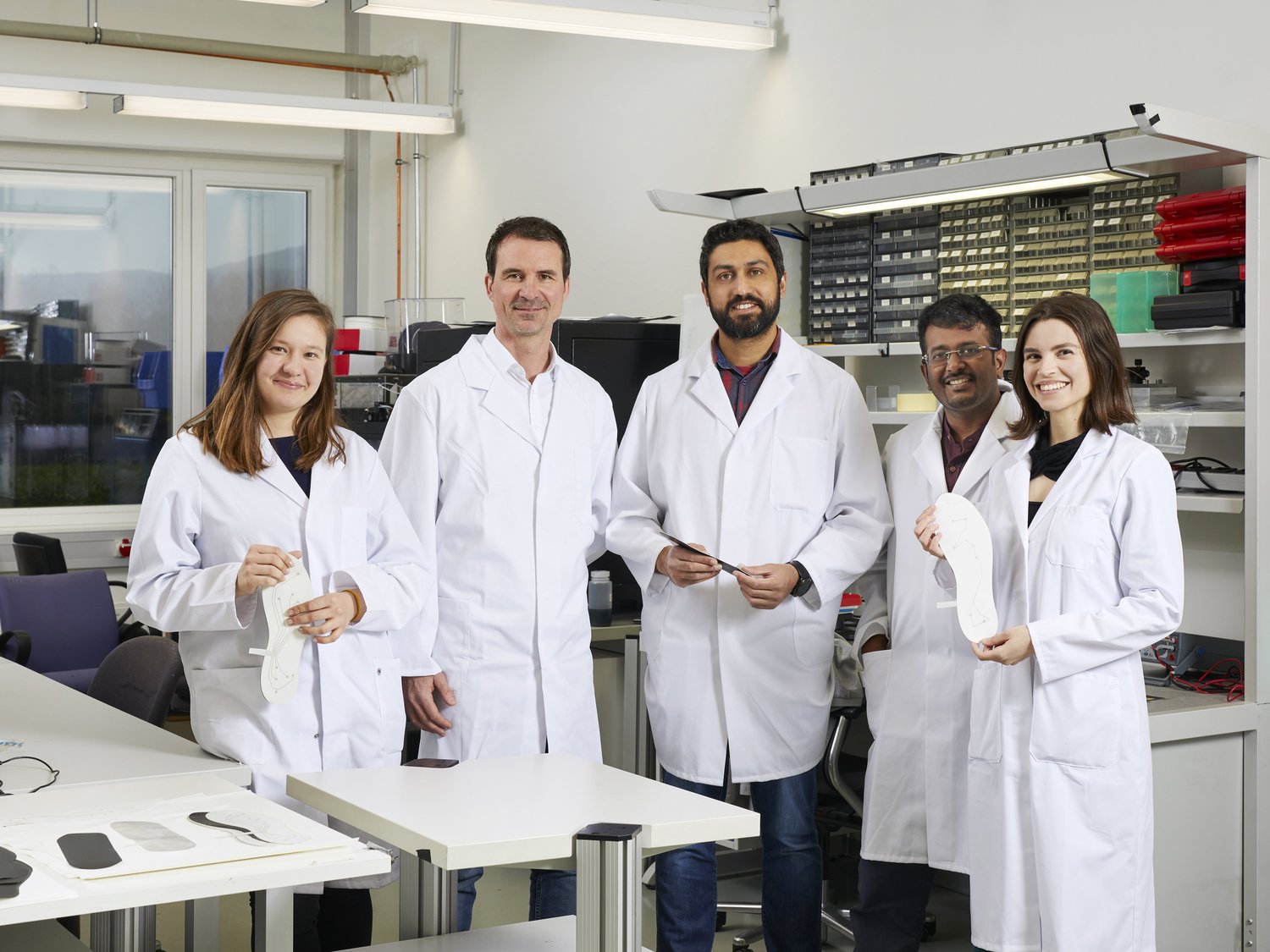 Tutku and four other researchers in a lab. Two are holding prototypes of intelligent shoe soles. 