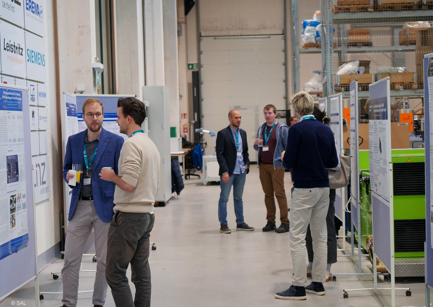 Menschen stehen in einer Fabrik vor wissenschaftlichen Postern auf Pinnwänden