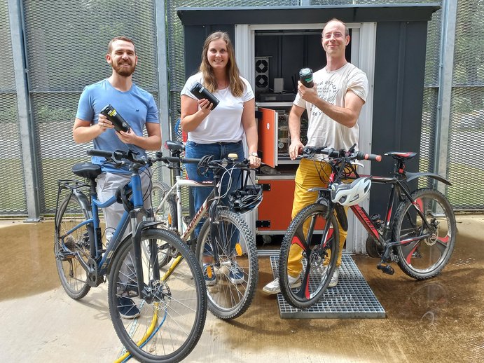 3 Mitarbeiter halten Fahrrad und Batterie zum elektrischen Antrieb des Rades; Ladestation im Hintergrund