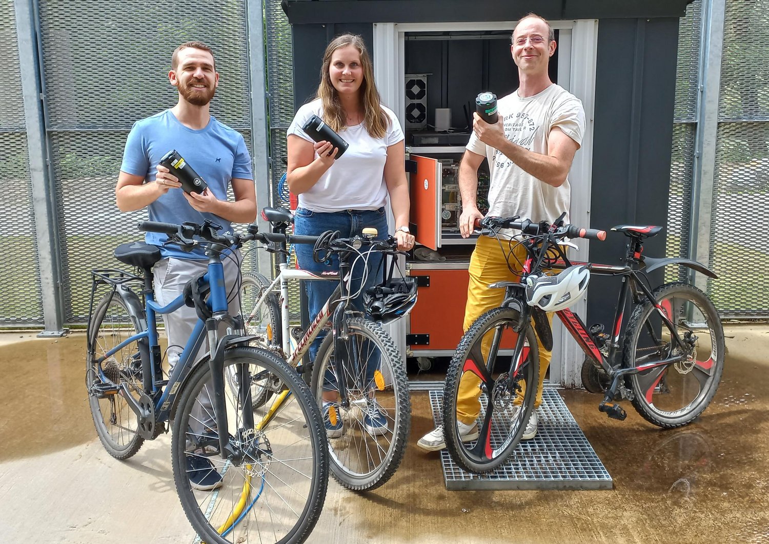 3 Mitarbeiter halten Fahrrad und Batterie zum elektrischen Antrieb des Rades; Ladestation im Hintergrund