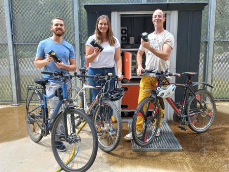 3 Mitarbeiter halten Fahrrad und Batterie zum elektrischen Antrieb des Rades; Ladestation im Hintergrund