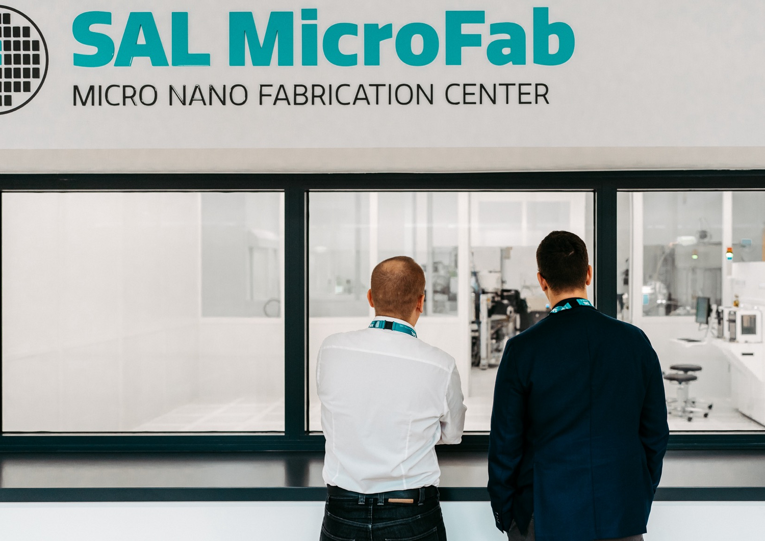 Two men stand in front of a large window and look into a research clean room with technical equipment